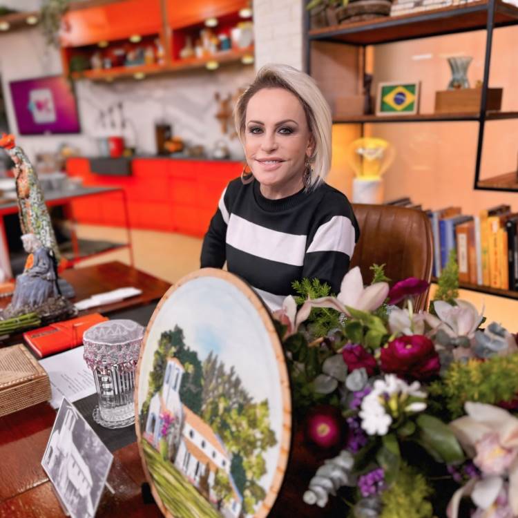 A imagem mostra uma pessoa sentada em um ambiente decorado. Ela tem cabelo loiro e veste uma blusa listrada em preto e branco. Na frente dela, há um arranjo de flores e uma pintura circular de uma paisagem com uma casa. Ao fundo, há prateleiras com livros e objetos, incluindo uma bandeira do Brasil. O ambiente parece ser acolhedor e bem decorado, com tons quentes e detalhes em vermelho.