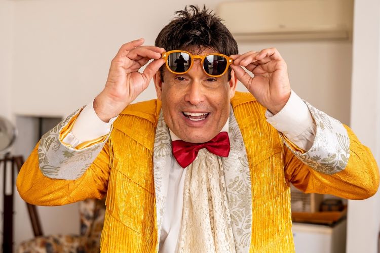 Sérgio Mallandro, humorista e apresentador brasileiro, veste um terno amarelo vibrante com detalhes brancos e uma gravata borboleta vermelha. Ele segura óculos de sol amarelos, exibindo seu característico sorriso jovial e carismático. A imagem destaca seu estilo irreverente e colorido, típico de sua personalidade cômica.