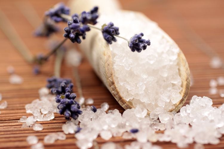 A imagem mostra um recipiente de madeira contendo sal grosso, com alguns cristais espalhados ao redor sobre uma superfície de madeira. Raminhos de lavanda estão dispostos sobre o sal, adicionando um toque de cor roxa e sugerindo uma combinação aromática e relaxante. A cena evoca uma sensação de tranquilidade e bem-estar.