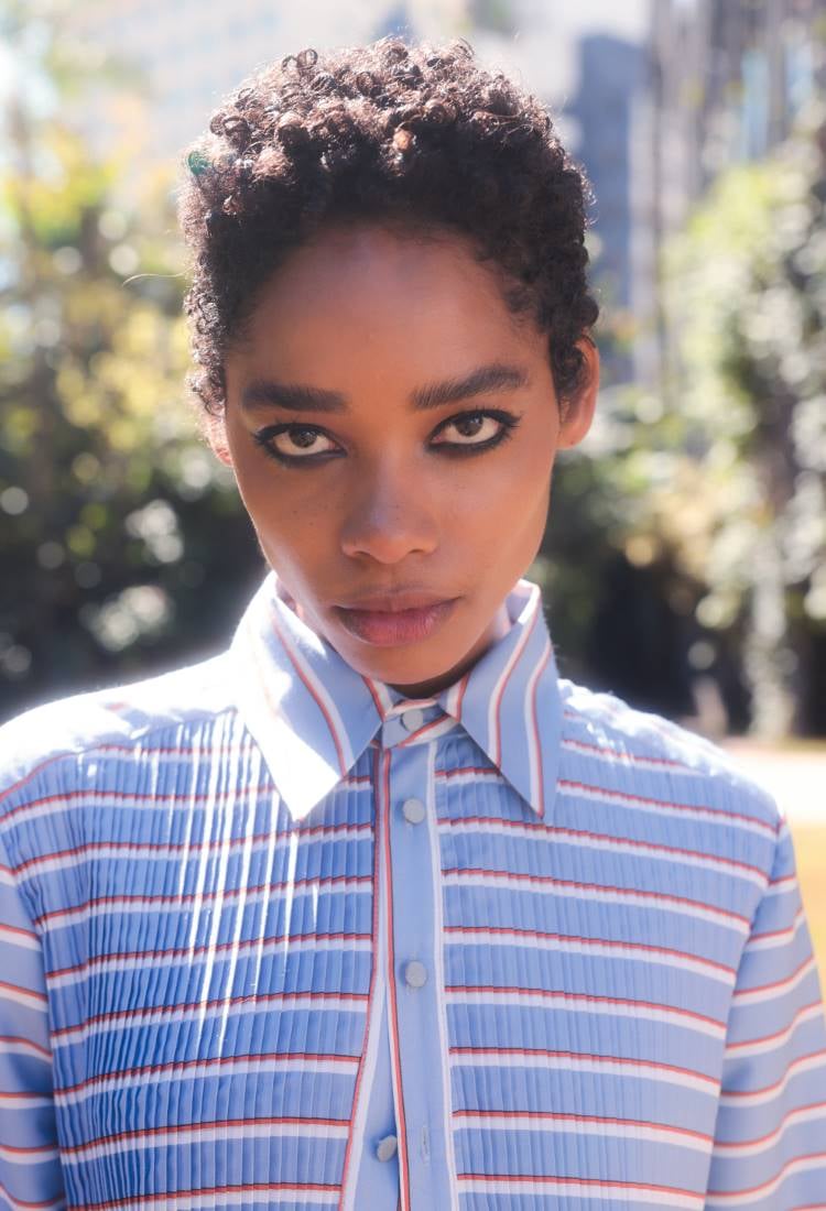 Outra modelo do desfile da Neriage patrocinado pela Eudora está ao ar livre, usando uma camisa azul clara com listras vermelhas e brancas. Seu cabelo é curto e encaracolado, e ela olha diretamente para a câmera com uma expressão séria. O fundo é desfocado e verde.