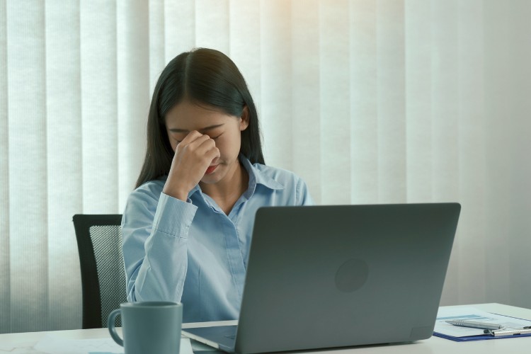 Mulher de pele clara sentada à mesa, usando camisa azul, em frente a um computador e com expressão triste 