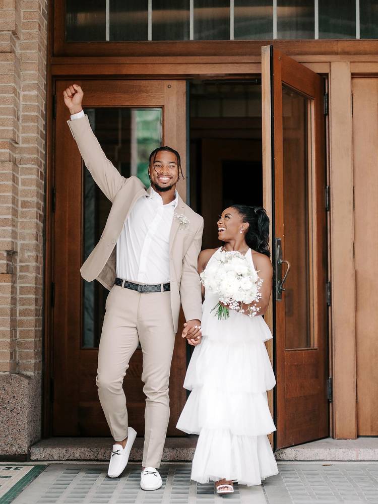 Jonathan Owens e Simone Biles saindo por porta de madeira casados. Ele com terno bege e camisa branca e ela com vestido de noiva 