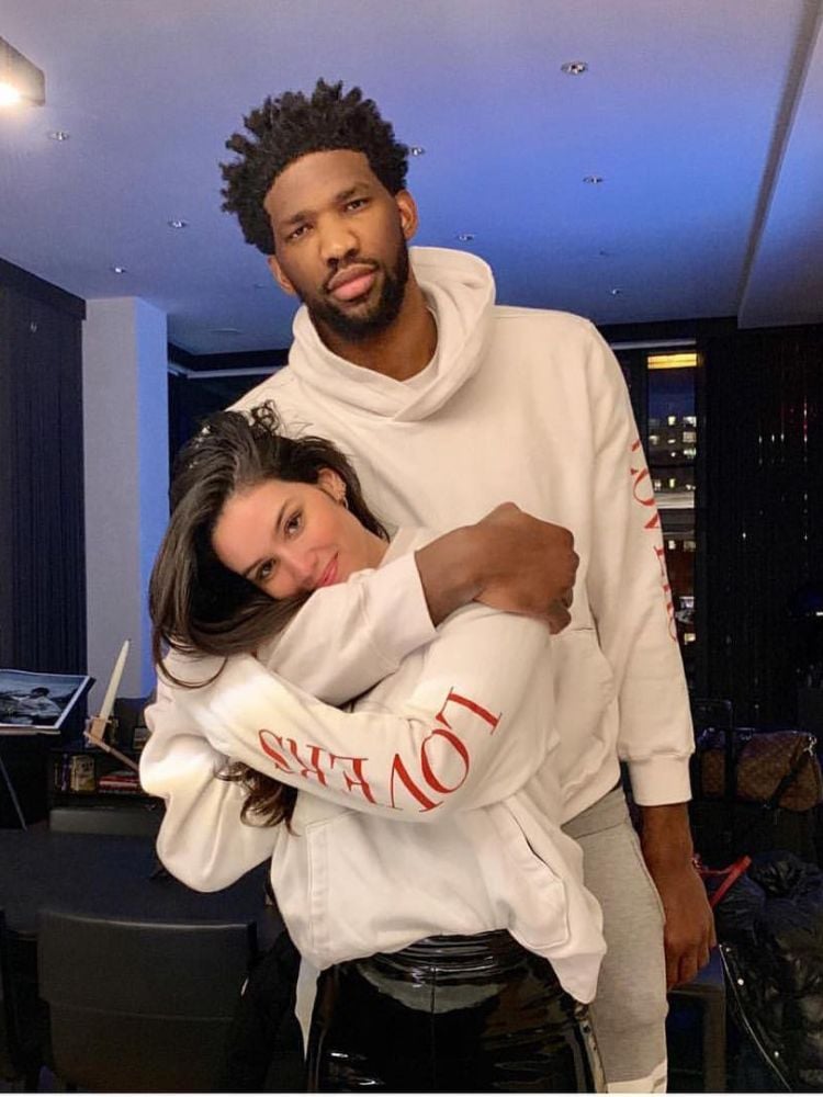 Joel Embiid e Anne de Paula abraçados, ambos usando moletom branco