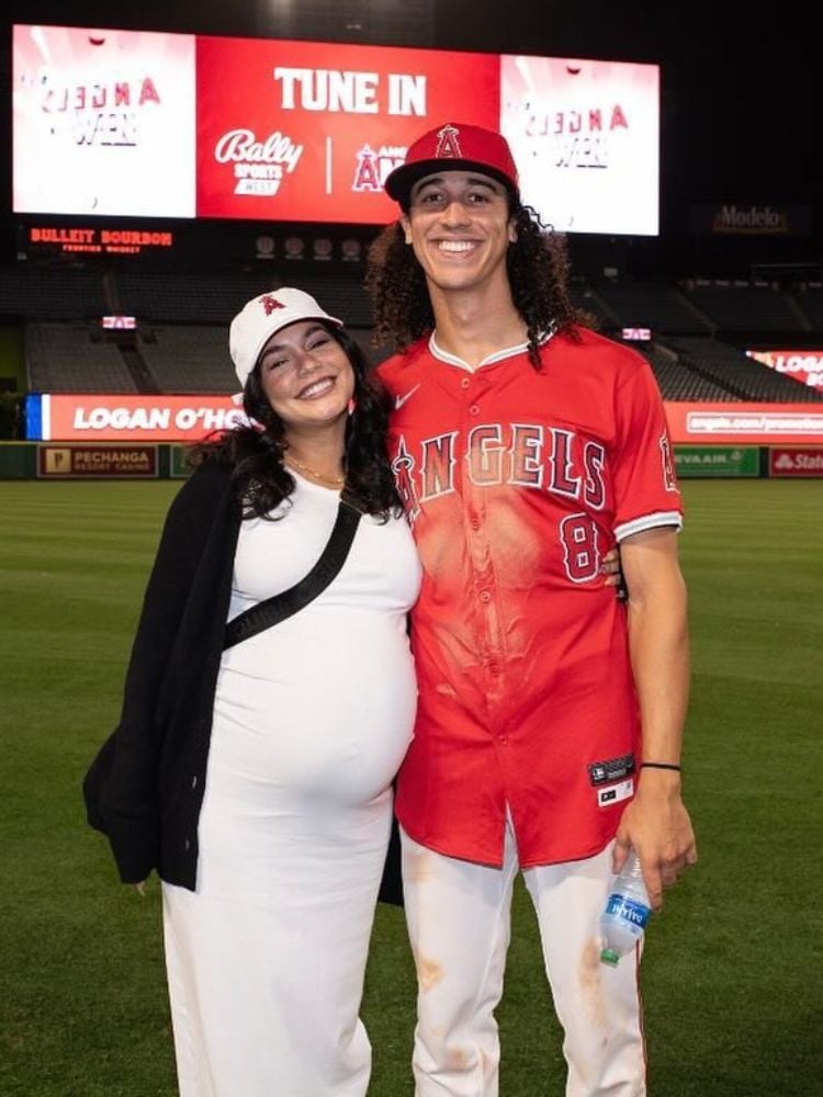 Vanessa Hudgens grávida usando vestido branco e casaco preto abraçada com seu marido Cole Tucker, que usando uniforme vermelho, boné e calça branca 