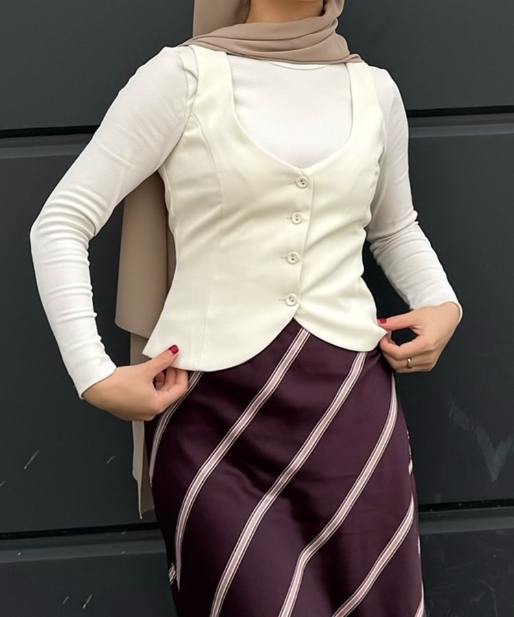 Uma mulher veste uma blusa branca de mangas compridas, colete branco ajustado e saia roxa com listras diagonais brancas. Ela usa um hijab bege. Este visual é um exemplo de moda modesta.