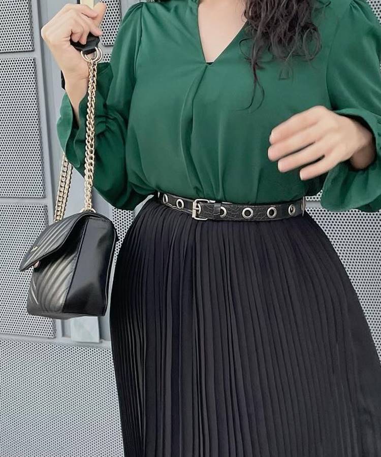 Uma mulher usa uma blusa verde de mangas longas, saia preta plissada e cinto preto com fivelas. Ela segura uma bolsa preta com corrente dourada. Este look elegante representa a moda modesta.