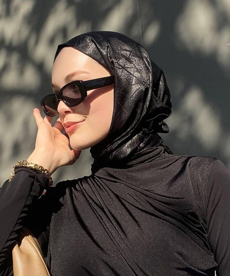 Uma mulher está em um ambiente externo, sob luz natural. Ela veste um hijab preto brilhante e uma blusa preta de mangas compridas. Usa óculos de sol grandes e retangulares. No pulso, há uma pulseira dourada. Este visual elegante e discreto é um exemplo de moda modesta.