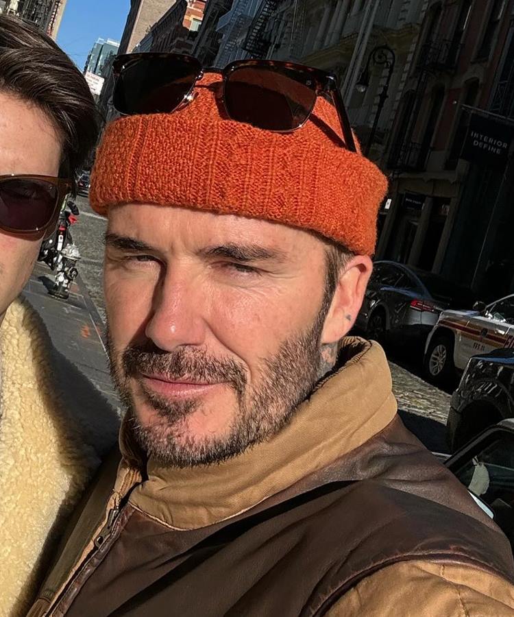 A imagem mostra David Beckham com barba e cabelo curto, usando um gorro laranja. Ele tem óculos de sol apoiados no gorro. Está vestindo uma jaqueta marrom e está em uma rua urbana, com prédios e carros ao fundo. A luz do sol ilumina seu rosto.