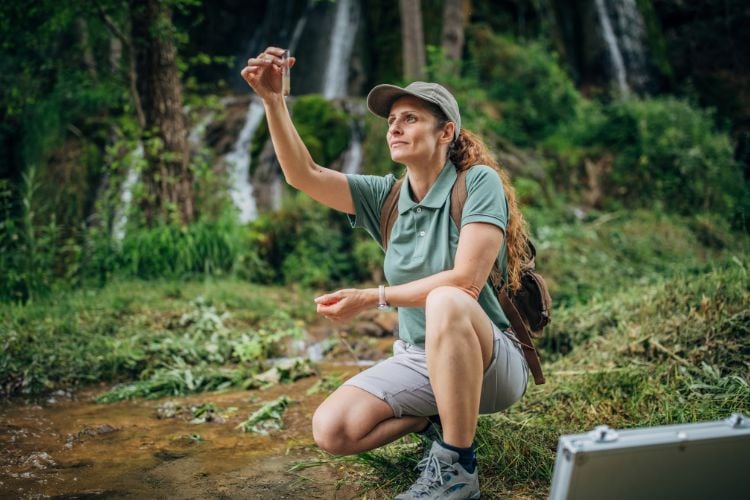 Mulher de pele clara usando blusa verde e bermuda cinza abaixada em floresta segurando tubo de coleta