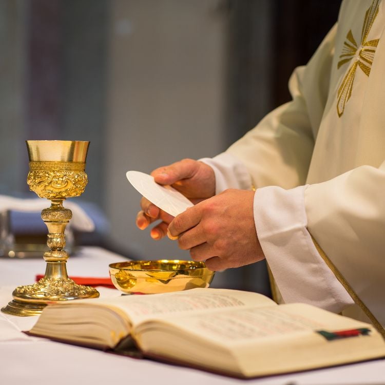 A imagem mostra um padre segurando uma hóstia branca em preparação para a Eucaristia. Ele veste uma batina branca com detalhes dourados. Na mesa, há um cálice dourado, uma patena e um livro aberto, provavelmente um missal. A cena ocorre durante uma missa.