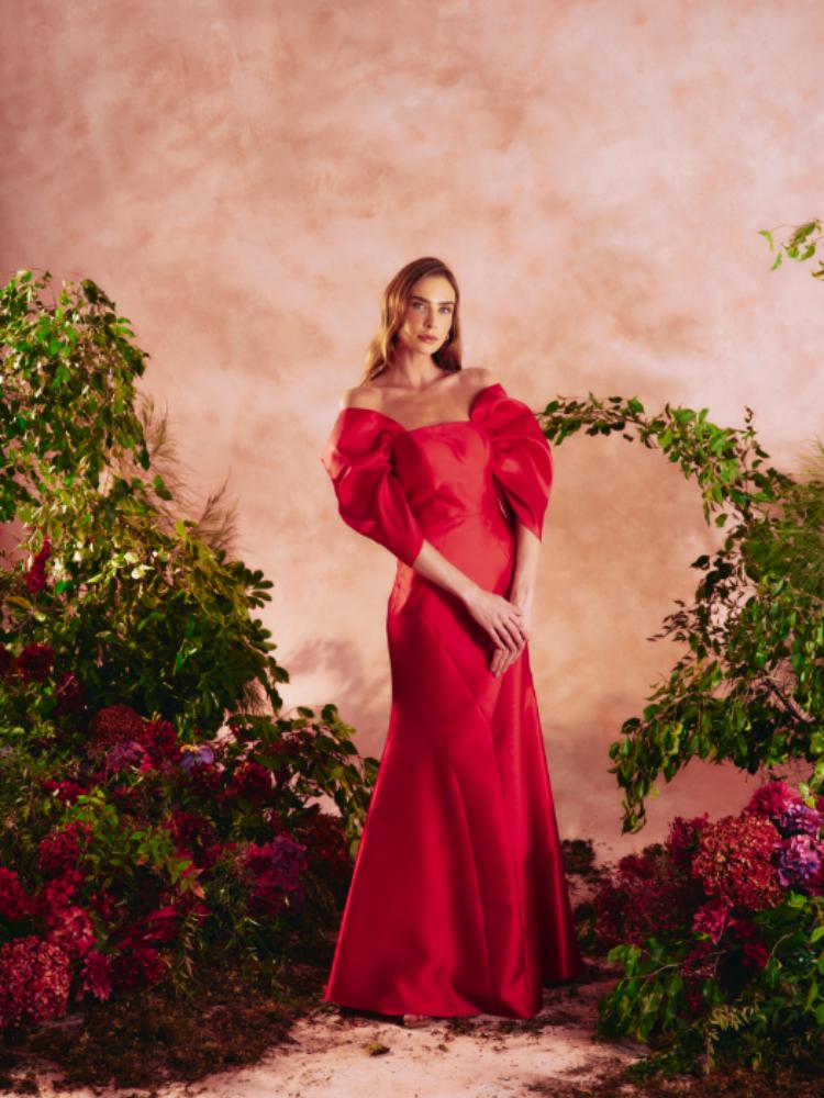 Mulher loira de pele clara usando vestido longo vermelho com manga bufante em paisagem de fundo bege com flores