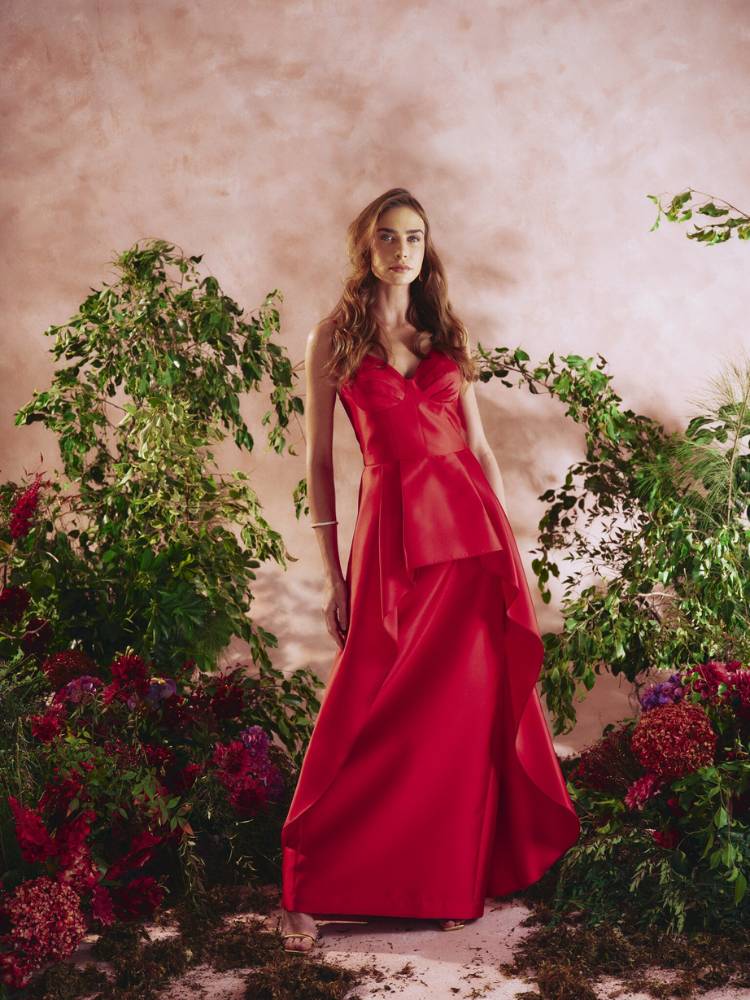 Mulher de pele clara usando vestido longo vermelho de alças com babado em fundo bege com flores ao redor