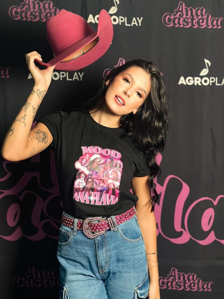 Ana Castela usando chapéu rosa, blusa preta com desenho rosa, cinto rosa com fivela com brilhos e calça jeans