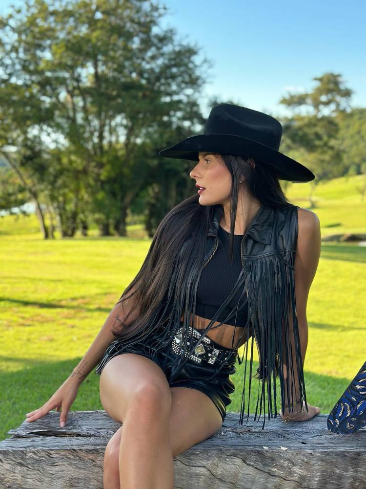 Ana Castela sentada em mureta em jardim usando top preto, colete regata preto com franja, chapéu preto, shorts preto e cinto preto com fivela prateada