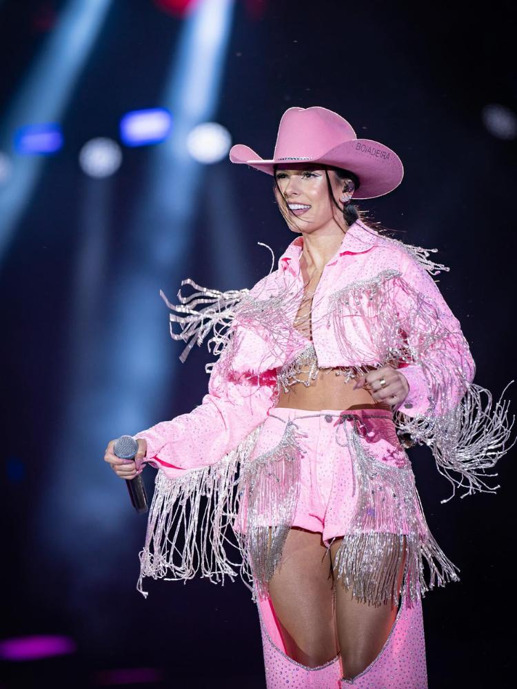 Ana Castela usando look rosa, com chapéu, top, jaqueta e calça com recorte de shorts com franjas