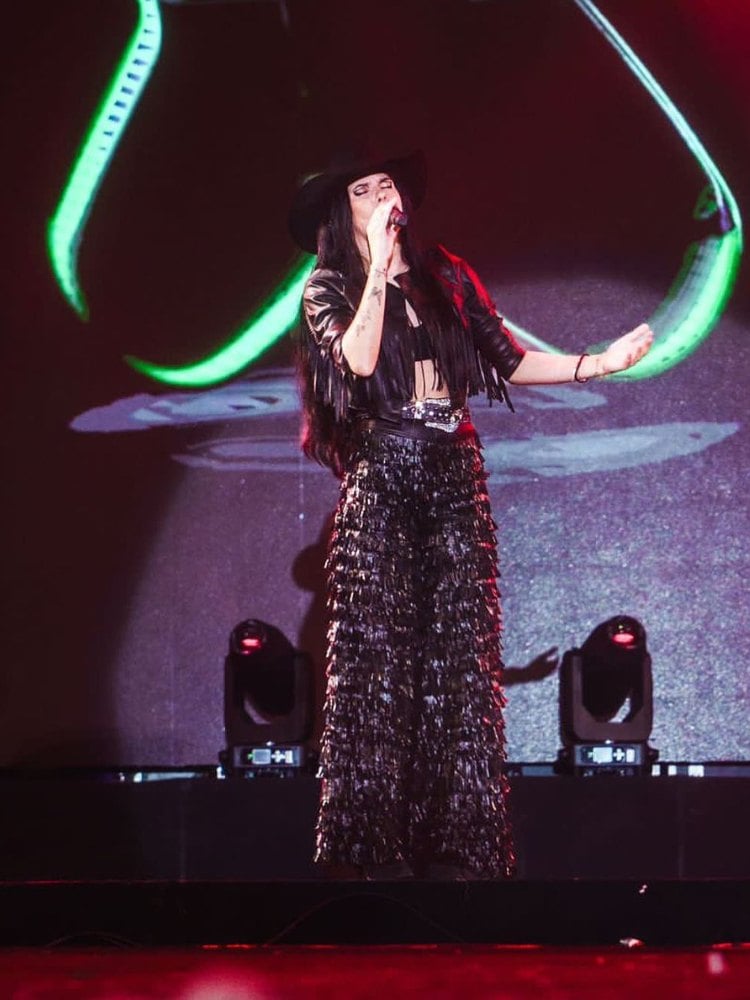 Ana Castela no palco usando jaqueta preta de couro com franjas, top preto, calça preta com franjas e chapéu preto