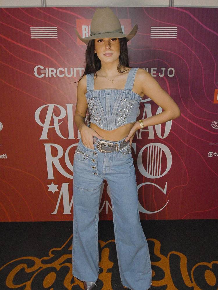 Ana Castela usando look all jeans, com cropped e calças com brilho, cinto marrom e chapéu bege