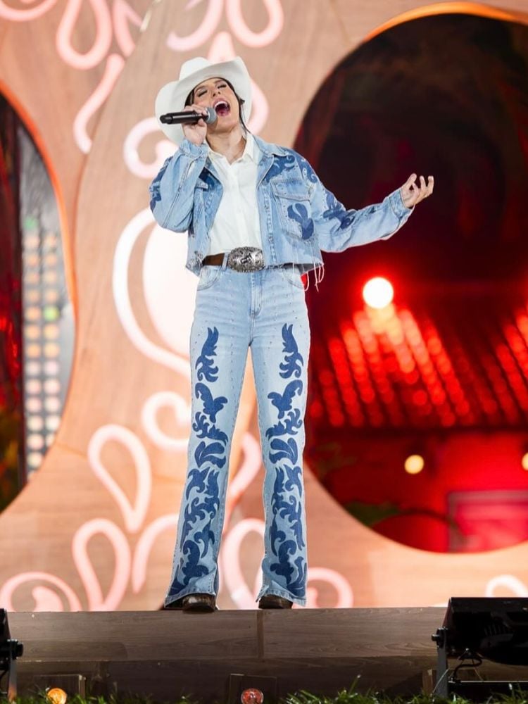 Ana Castela usando camisa branca, jaqueta jeans com bordados jeans escuros, cinto marrom, calça jeans no mesmo estilo da jaqueta, chapéu branco e bota marrom