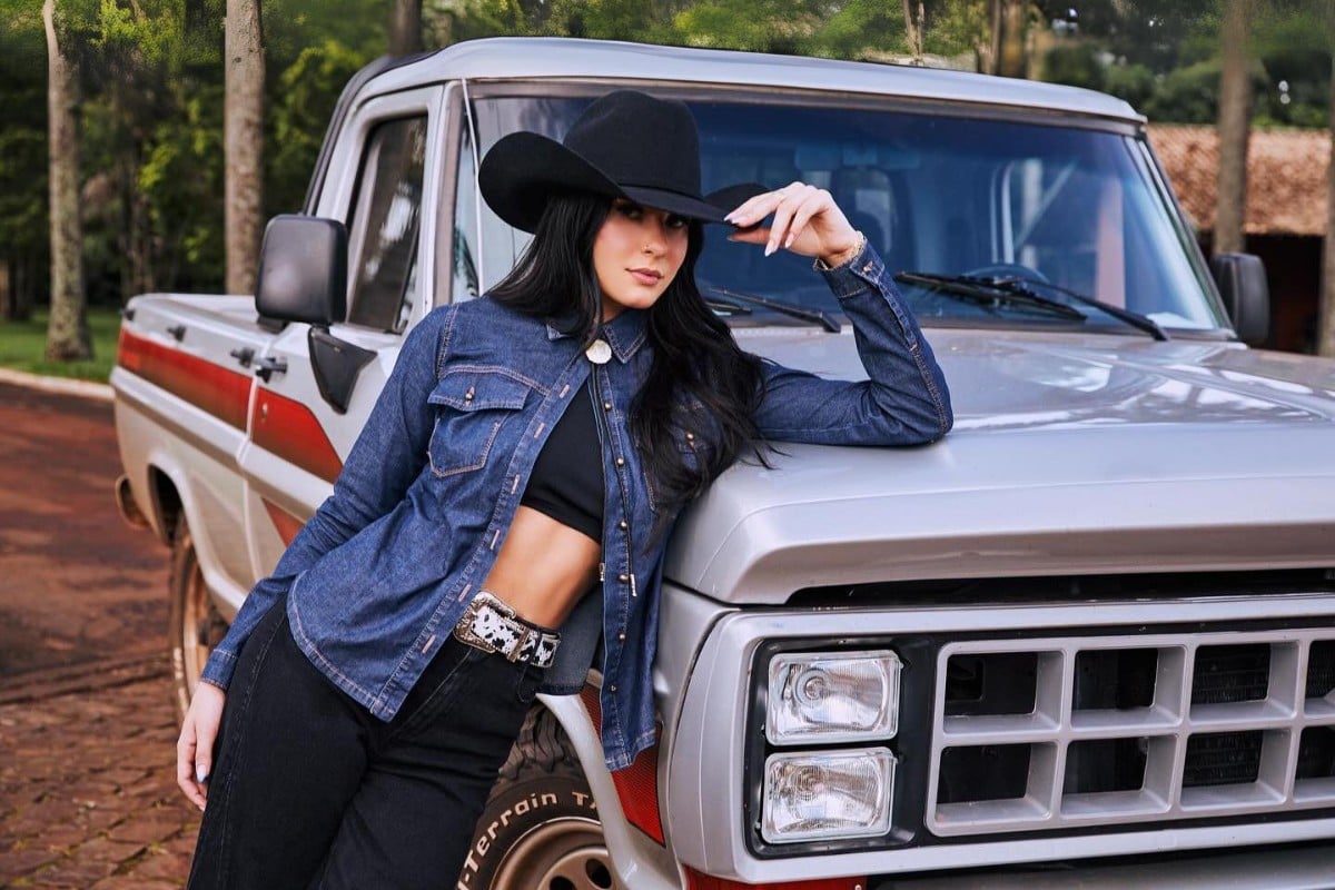 Foto de Ana Castela debruçada sobre caminhonete prateada, usando chapéu preto, top preto, jaqueta jeans e calça preta