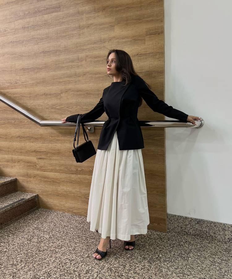 Em um ambiente interno, uma mulher veste um blazer preto ajustado e uma saia longa branca plissada. Ela segura uma bolsa preta e usa sandálias pretas. Este look representa a moda modesta.