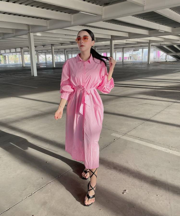 Em um estacionamento, uma mulher veste um vestido longo rosa com mangas bufantes e cinto na cintura, combinando com sandálias pretas de tiras. Ela usa óculos de sol laranja. Este visual é um exemplo de moda modesta.