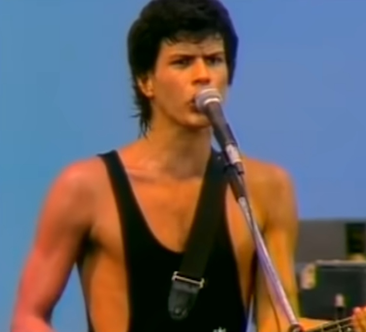 Paulo Ricardo, vocalista da banda RPM, durante uma performance ao vivo nos anos 80. Ele está usando uma regata preta e tocando guitarra enquanto canta ao microfone. A imagem captura a energia e o carisma que o tornaram um dos famosos anos 80 no cenário musical brasileiro.