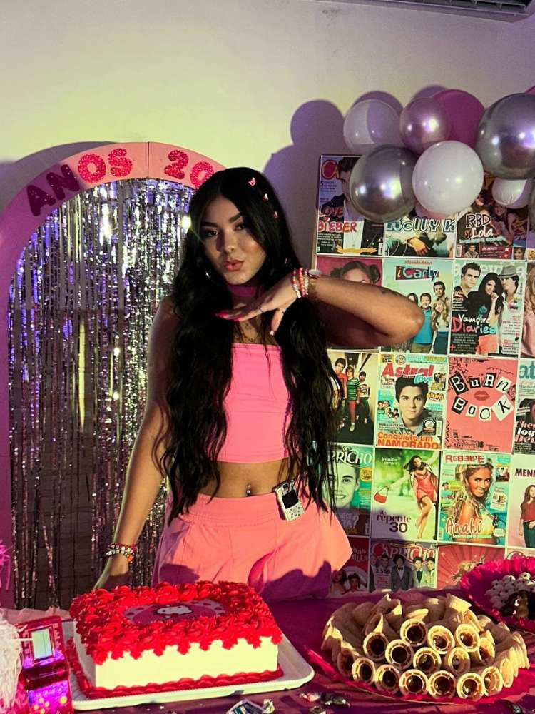 Mulher de pele morena com look rosa em festa dos anos 2000 decorada e com celular de flip preso na saia 