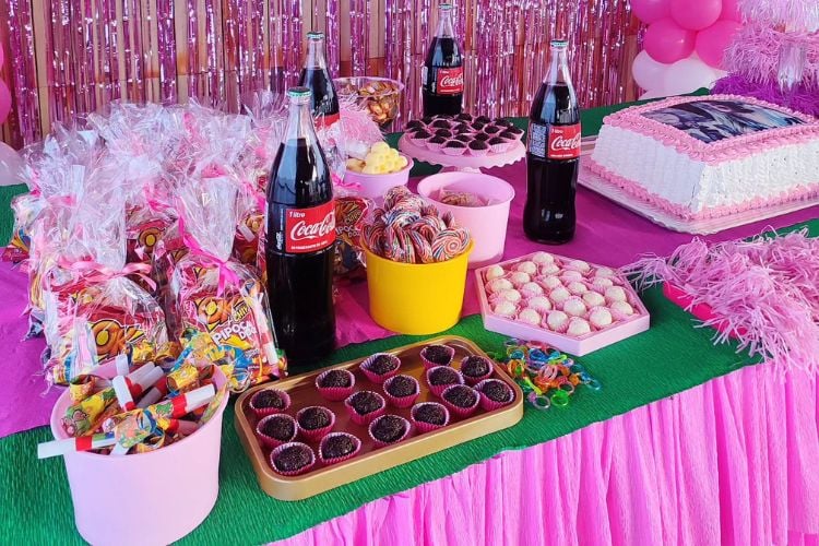 Mesa com papel crepom rosa, doces, refrigerantes e outras guloseimas 