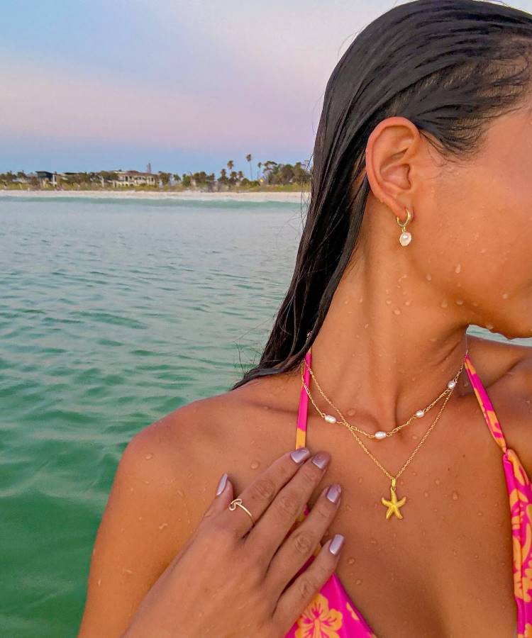 Uma mulher de cabelos molhados, usando um biquíni rosa com estampa floral, está no mar. Ela usa colares dourados e brincos de pérola, com a mão tocando o pescoço. Ao fundo, há uma praia com palmeiras. Fotos tumblr na praia frequentemente capturam momentos assim.