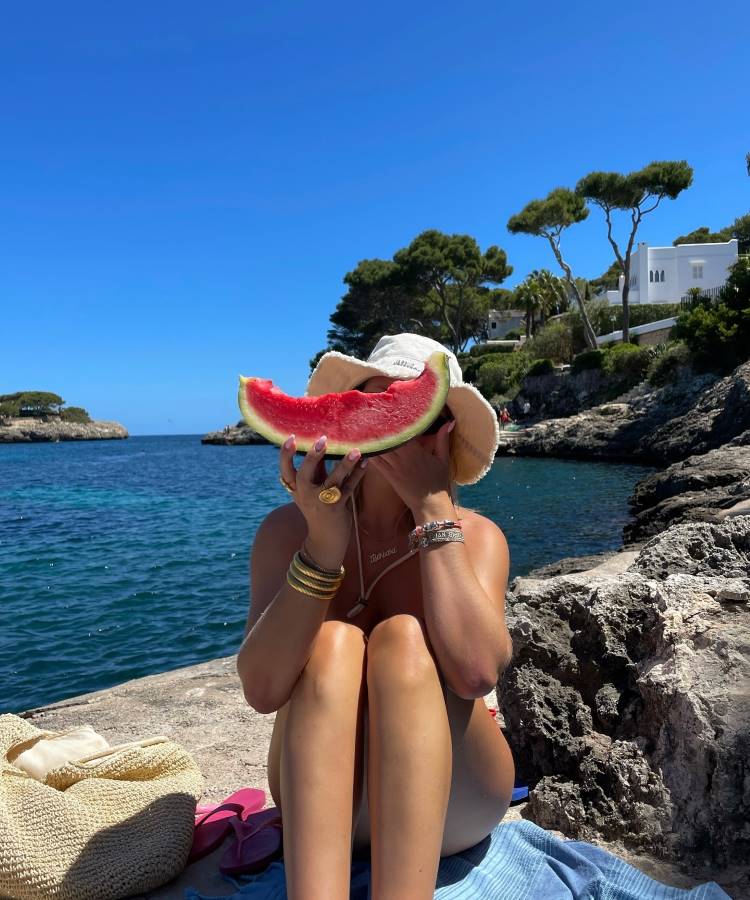 Uma mulher sentada em uma rocha à beira-mar, usando um chapéu de sol e segurando uma fatia de melancia na frente do rosto. Ela veste pulseiras douradas e tem uma bolsa de palha ao lado. Fotos tumblr na praia frequentemente mostram momentos descontraídos e divertidos como este.