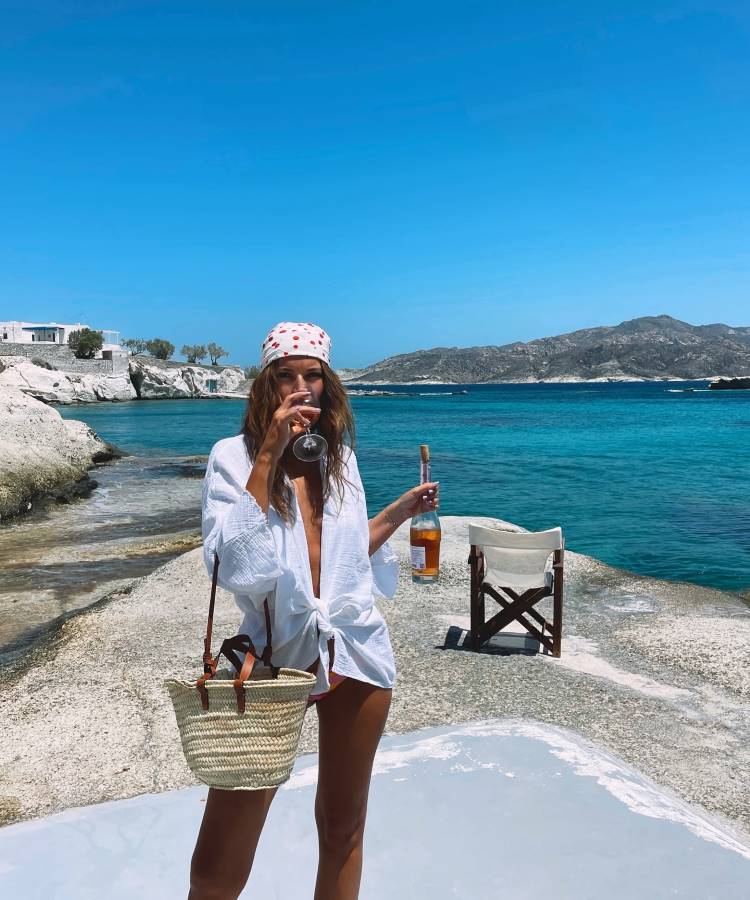 Uma mulher de cabelos longos e soltos, usando uma camisa branca e um lenço na cabeça, está em uma praia rochosa. Ela segura uma taça de vinho e uma garrafa, com uma bolsa de palha pendurada no ombro. Fotos tumblr na praia capturam momentos assim.