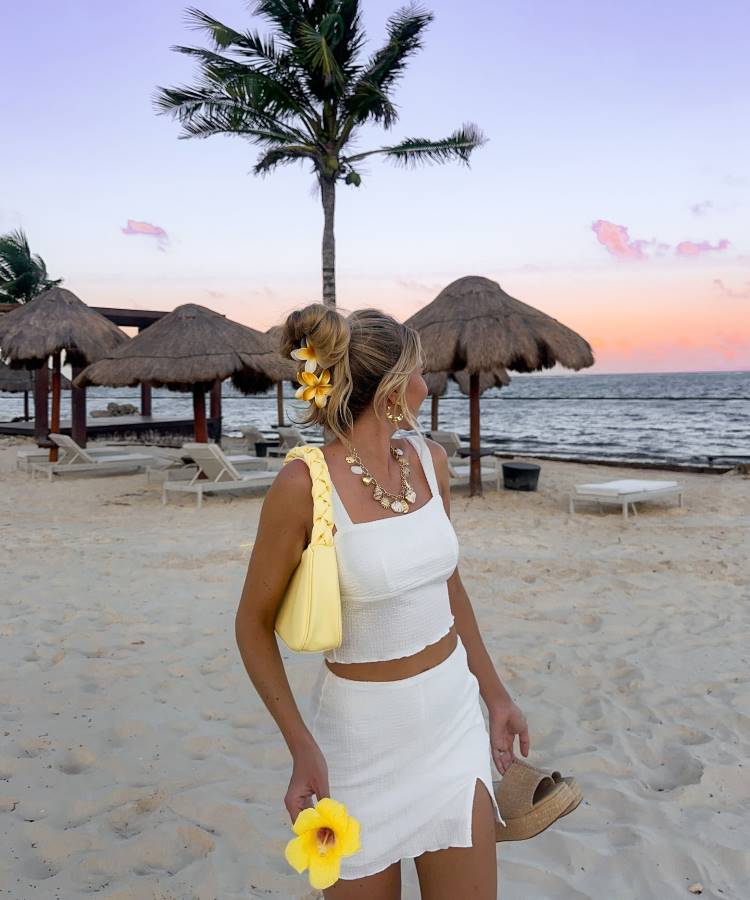 Uma mulher loira, com cabelo preso e decorado com flores amarelas, usa um conjunto branco de top e saia. Ela segura uma flor amarela e uma bolsa amarela, com palmeiras e cabanas de palha ao fundo, ao pôr do sol. Fotos tumblr na praia frequentemente mostram essa atmosfera.