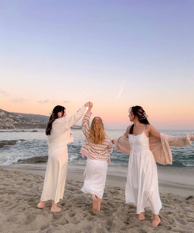 Três mulheres de costas, vestindo roupas claras e soltas, dançam na areia ao pôr do sol. Elas estão de mãos dadas, com o mar e o céu alaranjado ao fundo. Fotos tumblr na praia mostram momentos de alegria e amizade em cenários deslumbrantes.