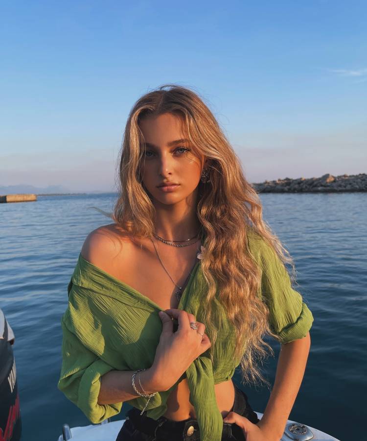 Uma mulher loira com cabelo ondulado, vestindo uma blusa verde amarrada na frente, está em um barco com o mar ao fundo. O céu está azul e o sol está se pondo, iluminando seu rosto.