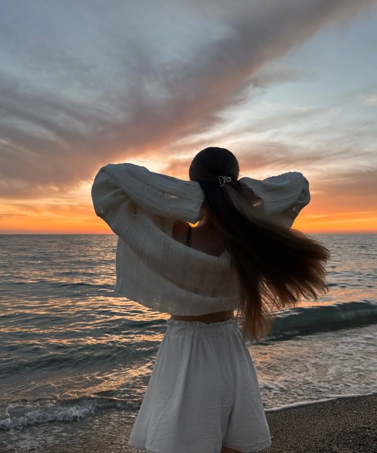 Uma mulher de cabelos longos, vestindo um conjunto branco de blusa e shorts, está de costas na praia ao pôr do sol. Ela segura o cabelo enquanto observa o mar e o céu alaranjado. Fotos tumblr na praia frequentemente mostram essa atmosfera serena e mágica.