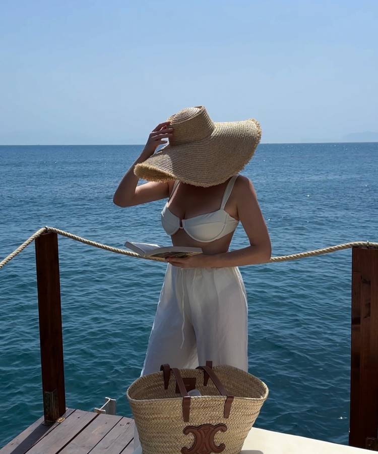 Uma mulher de biquíni branco e calça larga branca está em um deck de madeira, segurando um chapéu de palha grande que cobre seu rosto. Ela lê um livro, com uma bolsa de palha ao lado e o mar azul ao fundo. Fotos tumblr na praia capturam essa serenidade.