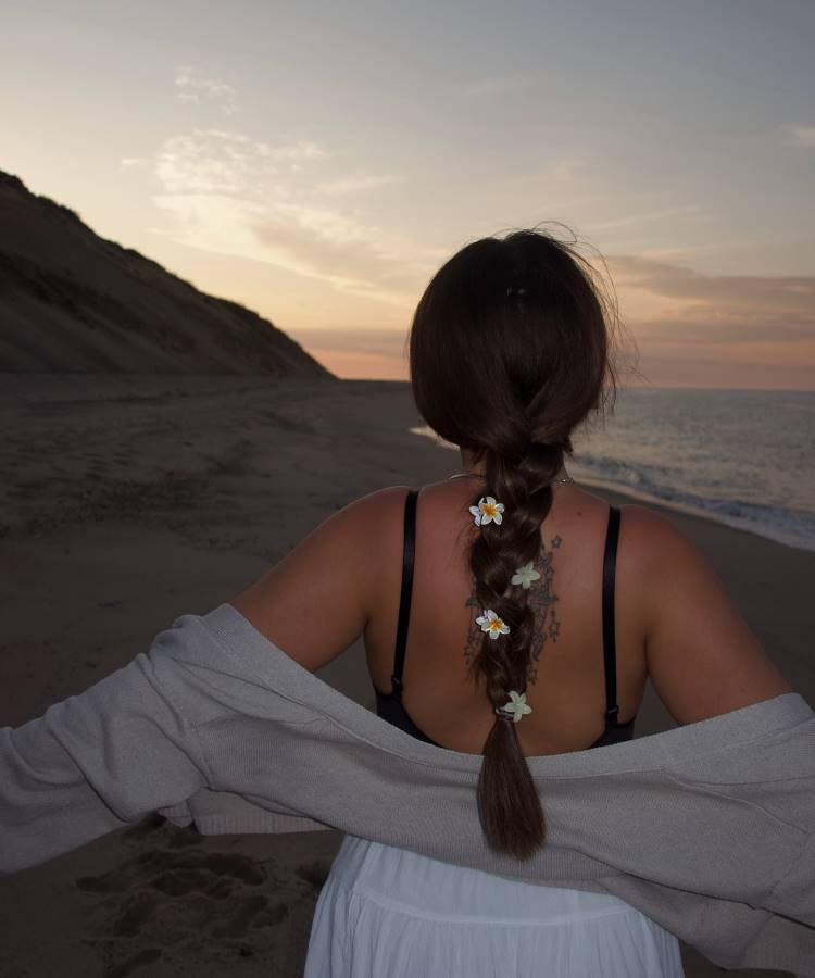 Uma mulher de costas, com uma trança decorada com flores brancas, está na praia ao pôr do sol. Ela usa um top preto e uma saia branca, com um casaco cinza caindo dos ombros. O mar e um penhasco estão ao fundo.