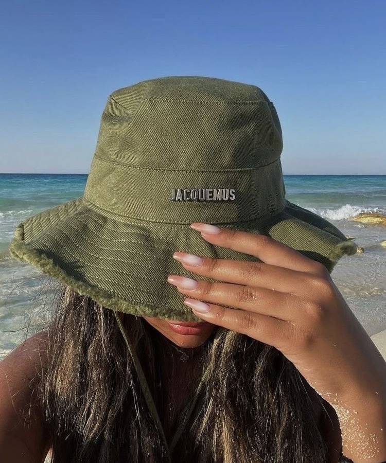 Uma mulher de cabelos longos e escuros, usando um chapéu verde, está na praia. Ela cobre parcialmente o rosto com a mão, exibindo unhas bem cuidadas. Ao fundo, o mar azul e o céu claro completam a cena. Fotos tumblr na praia mostram essa atmosfera relaxante.