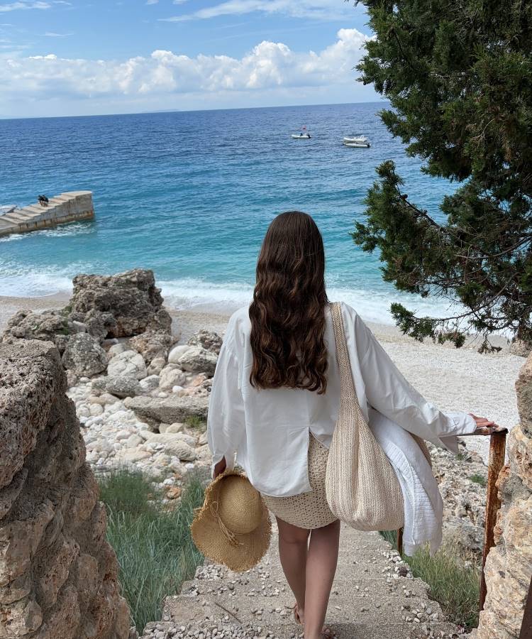 Uma mulher de cabelos longos e ondulados, vestindo uma camisa branca e uma saia de crochê, desce uma escada de pedra em direção a uma praia de águas azuis. Ela carrega um chapéu de palha e uma bolsa de crochê, com árvores e rochas ao redor.