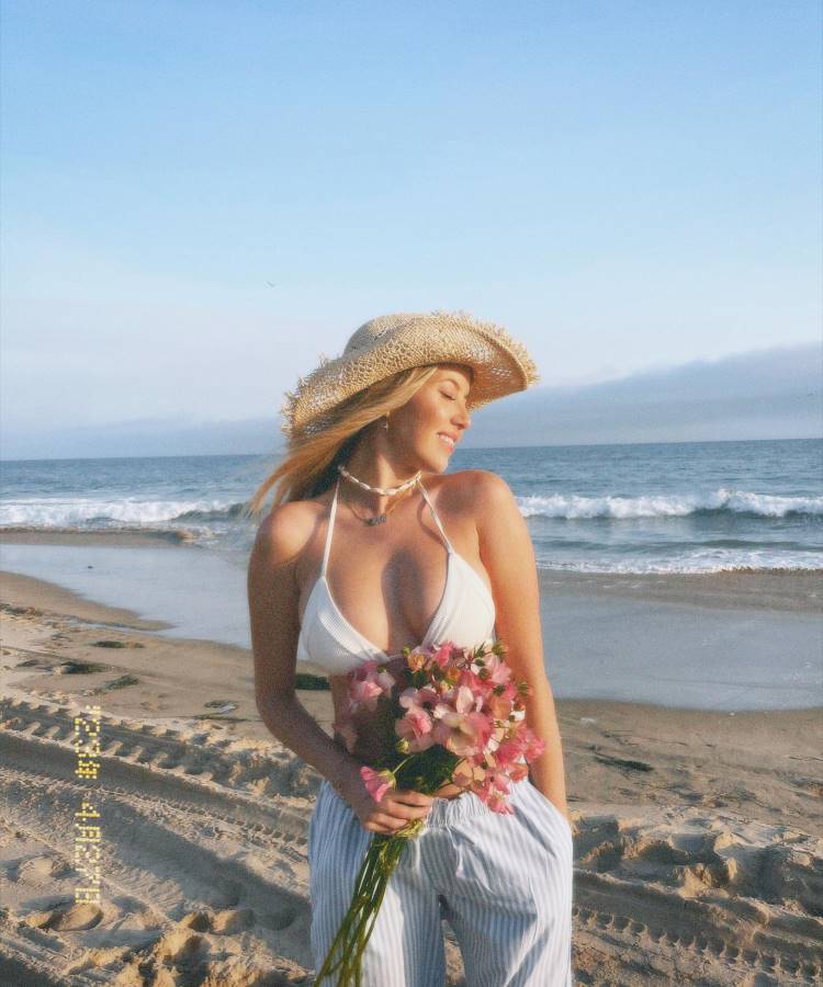 Uma mulher loira, usando um chapéu de palha e um top branco, está na praia segurando um buquê de flores cor-de-rosa. Ela veste calças listradas e sorri suavemente, com o mar e o céu azul ao fundo. Fotos tumblr na praia capturam essa serenidade.