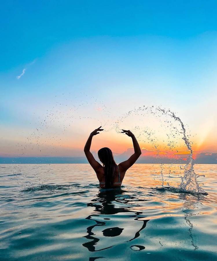 Uma mulher de costas, com cabelos longos e soltos, está no mar ao pôr do sol. Ela levanta os braços, criando um arco de água ao seu redor. O céu está azul e alaranjado. Fotos tumblr na praia frequentemente capturam momentos mágicos como este.