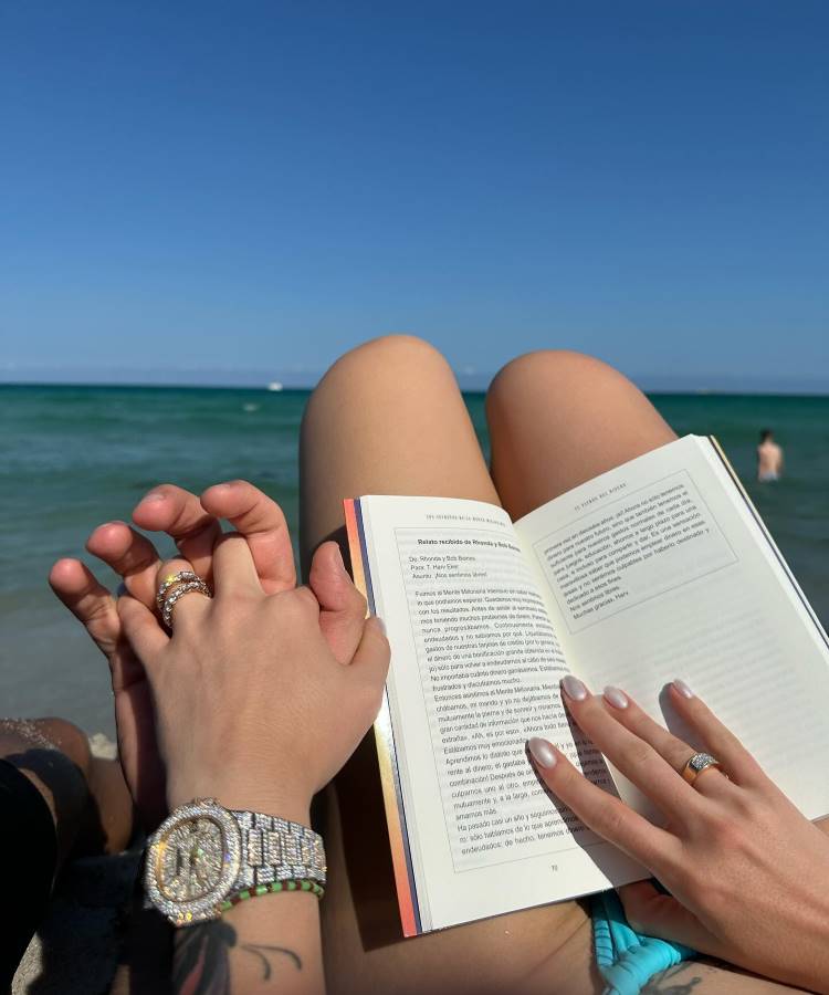 Uma mulher deitada na areia, usando um biquíni floral e shorts brancos, segura uma câmera rosa. Ela tem óculos de sol na cabeça e uma bolsa de praia ao lado. O mar e o céu nublado estão ao fundo. Fotos tumblr na praia mostram momentos relaxantes e descontraídos.