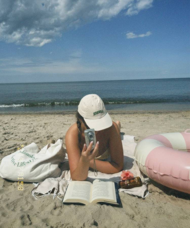 Uma mulher deitada na areia da praia, lendo um livro. Ela usa um boné branco e segura uma câmera. Ao seu lado, há uma bolsa de praia e um protetor solar. O mar e o céu azul com nuvens completam a cena.