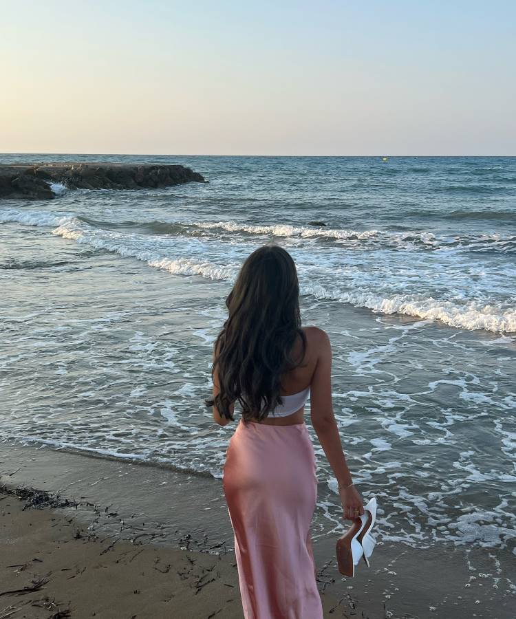 Uma mulher de cabelos longos e escuros, vestindo um top branco e uma saia longa rosa, caminha descalça na beira do mar. Ela segura sapatos brancos na mão, enquanto as ondas tocam suavemente a areia. O céu está claro, criando uma cena serena.