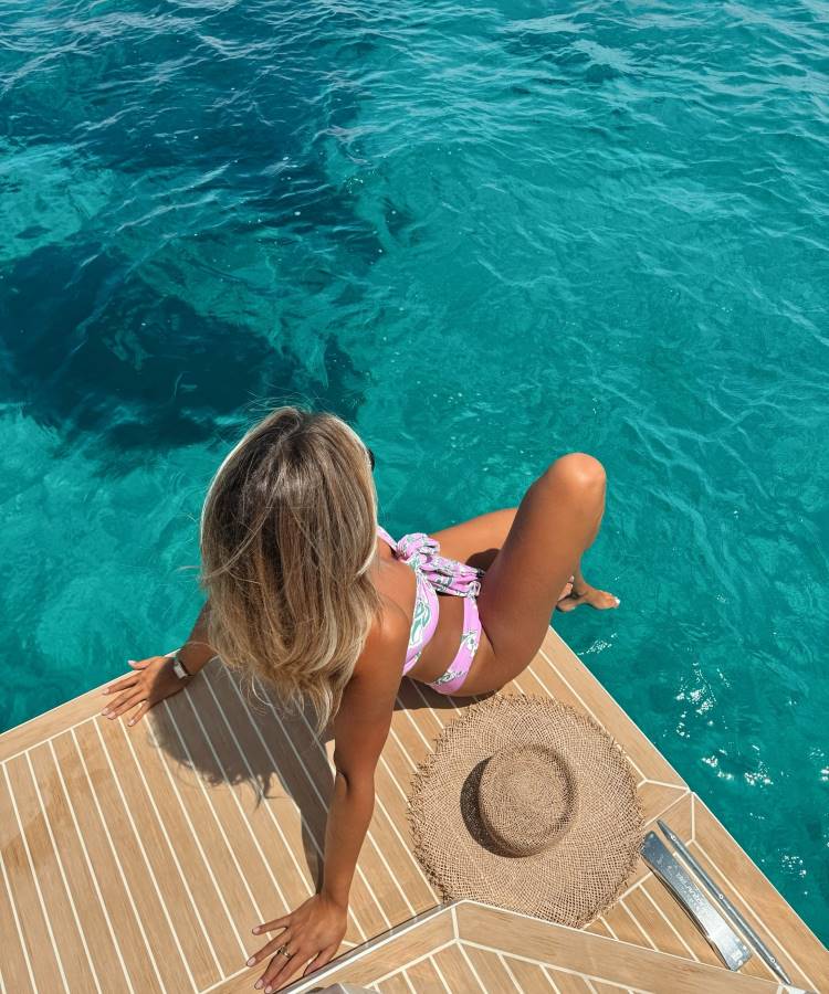 Uma mulher loira, de biquíni rosa com estampa floral, está sentada na borda de um barco de madeira sobre águas cristalinas. Ao lado dela, há um chapéu de palha. Fotos tumblr na praia frequentemente mostram momentos relaxantes como este, com o mar azul ao fundo.