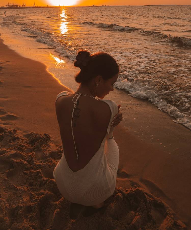 Uma mulher de cabelos presos, usando um vestido branco com as costas abertas, está ajoelhada na areia da praia ao pôr do sol. O mar e o céu alaranjado criam uma atmosfera serena e romântica, com a luz dourada refletindo na água.