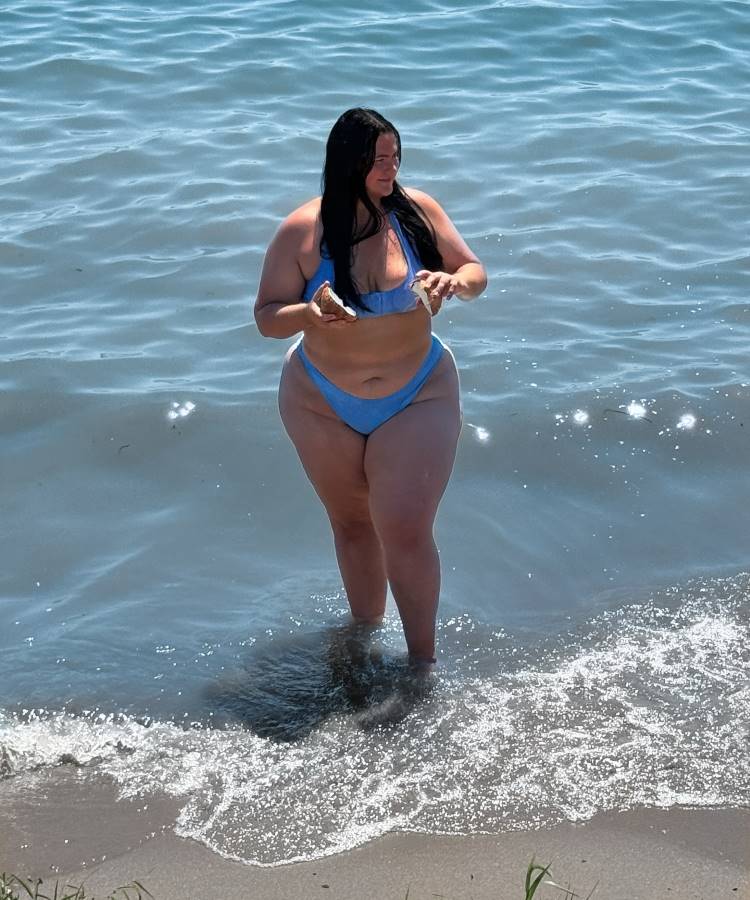 Uma mulher de cabelos longos e escuros, usando um biquíni azul, está na beira do mar. Ela segura um coco partido ao meio, com a água tocando seus pés. Fotos tumblr na praia mostram momentos descontraídos e naturais como este.