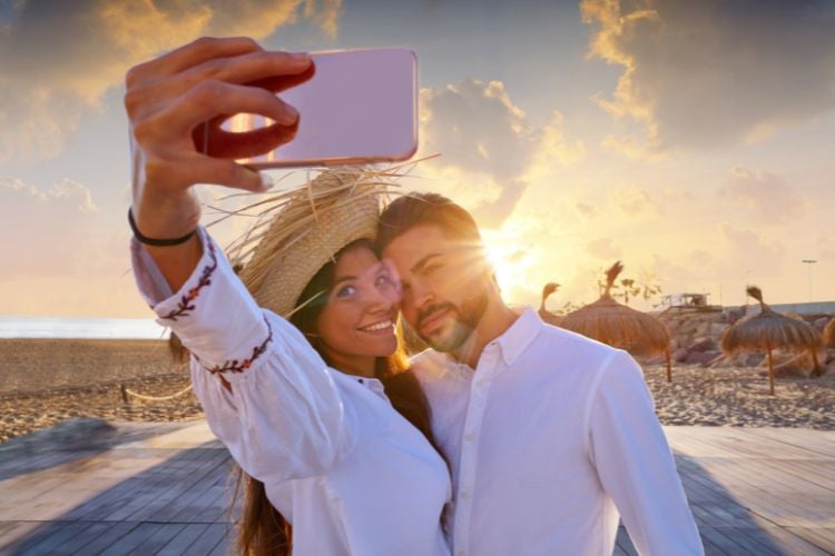 Um casal tira uma selfie na praia ao pôr do sol. Ambos estão sorrindo, a mulher usa um chapéu de palha e o homem está encostado nela. Eles vestem camisas brancas.