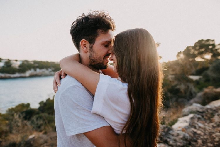 Um casal se abraça em um cenário natural ao pôr do sol. Eles estão de frente um para o outro, sorrindo, e vestem roupas casuais. Ao fundo, há árvores e um corpo d'água.