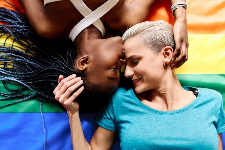 Duas mulheres estão deitadas de costas em uma bandeira do arco-íris, símbolo do orgulho LGBTQ+. Elas estão de frente uma para a outra, com os rostos próximos e sorrindo. Uma delas tem cabelo curto e loiro, e a outra tem cabelo longo e trançado. Elas estão em um momento de carinho e intimidade.